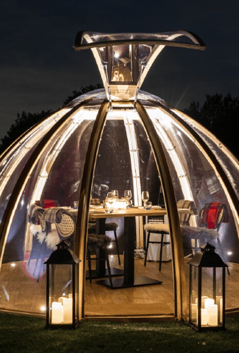 A cozy glass dome tent is dimly lit with fairy lights at night in the gardens of Stirrups Hotel. Inside, a dining table set elegantly for dinner. Two lanterns with candles frame the entrance.