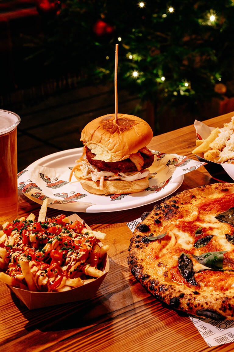 A table with a wood-fired pizza topped with basil, a burger on a plate, loaded fries, churros with dip, and a foamy glass of beer. Warm, inviting ambiance.