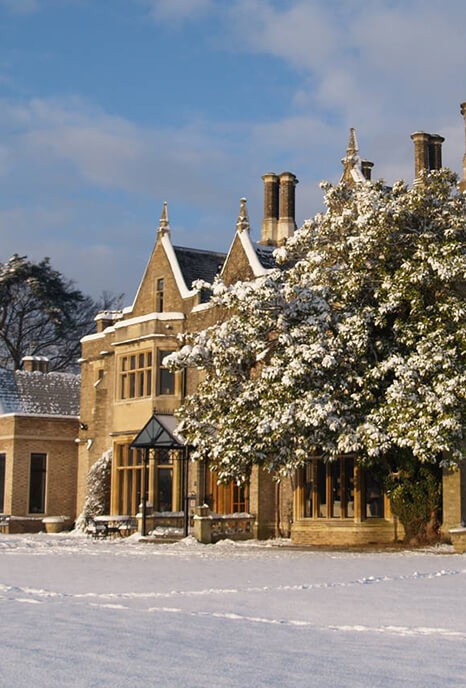 The Foxhills Resort and Spa, a grand stone mansion with tall chimneys and gabled roofs, covered in snow, surrounded by winter trees under a blue sky, conveying a serene winter scene.