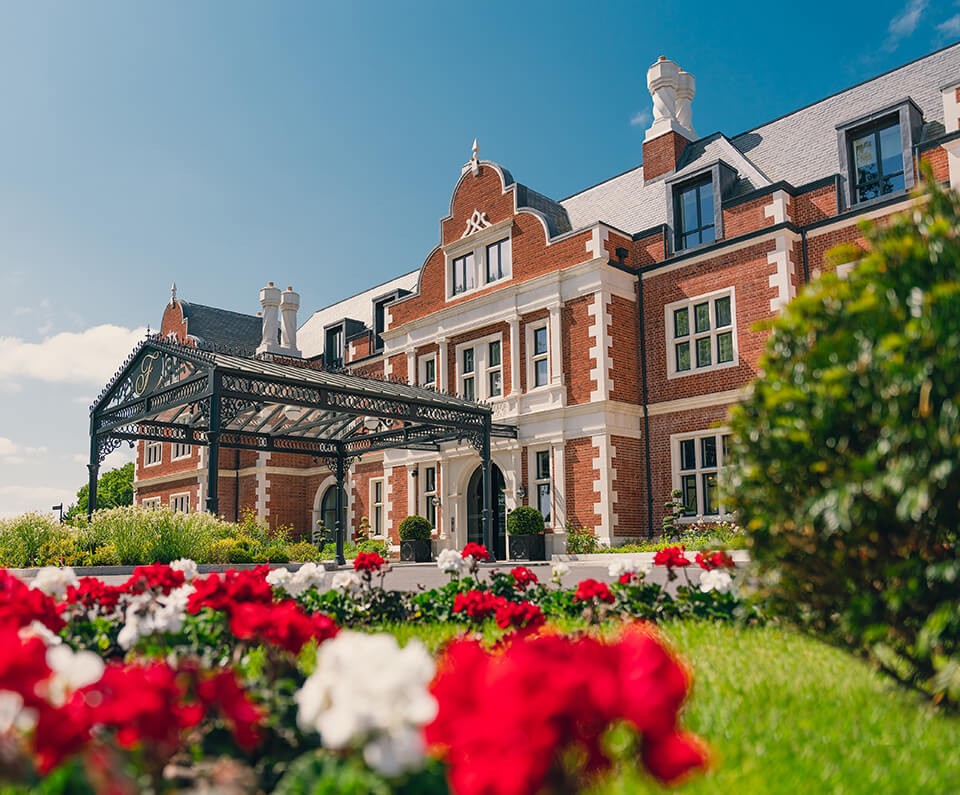 The Fairmont Hotel, a grand red-brick Victorian building with ornate black iron entrance stands under a clear blue sky, surrounded by vibrant red and white flowers.