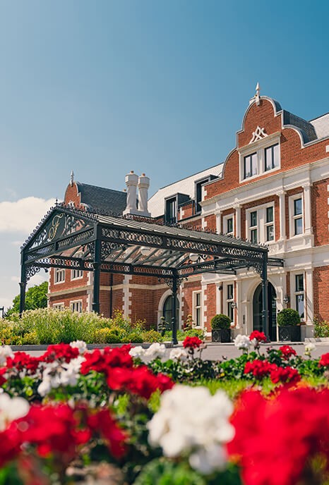 The Fairmont Hotel, a grand red-brick Victorian building with ornate black iron entrance stands under a clear blue sky, surrounded by vibrant red and white flowers.
