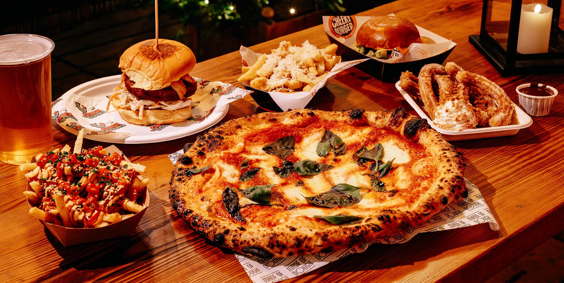 A table with a wood-fired pizza topped with basil, a burger on a plate, loaded fries, churros with dip, and a foamy glass of beer. Warm, inviting ambiance.