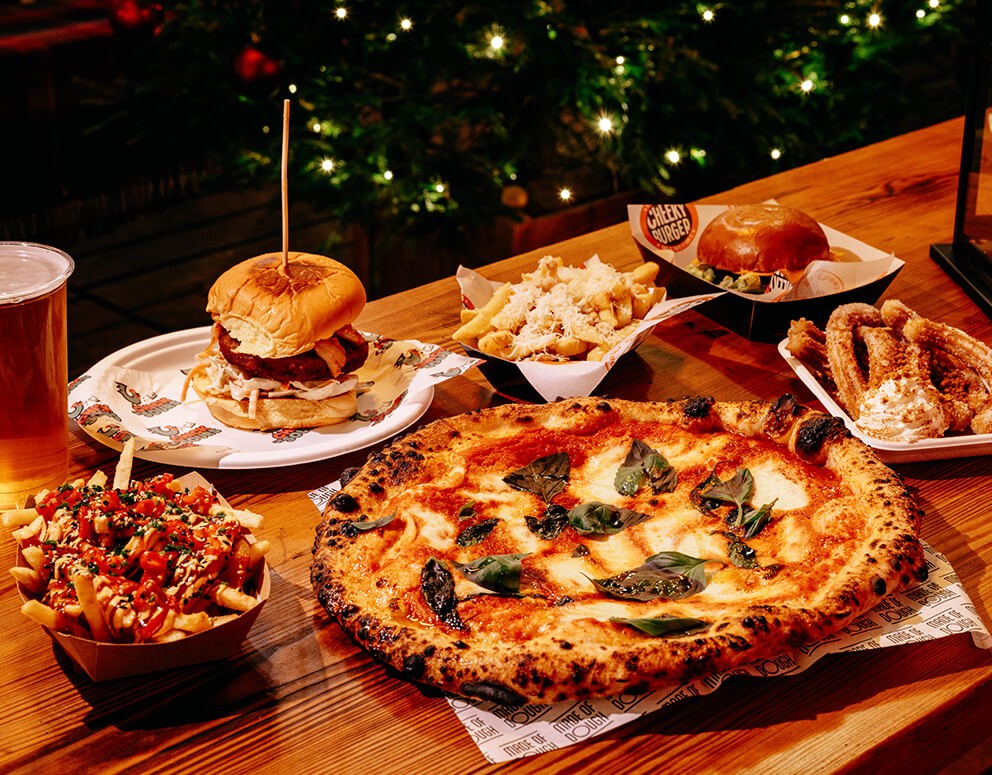 A table with a wood-fired pizza topped with basil, a burger on a plate, loaded fries, churros with dip, and a foamy glass of beer. Warm, inviting ambiance.