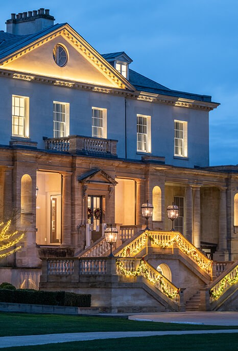 The Langley, an elegant mansion exterior at dusk with warm lights illuminating the facade and steps. A tree adorned with glowing fairy lights enhances the inviting ambiance.