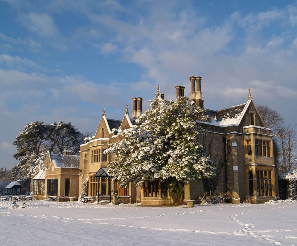 The Foxhills Resort and Spa, a grand stone mansion with tall chimneys and gabled roofs, covered in snow, surrounded by winter trees under a blue sky, conveying a serene winter scene.