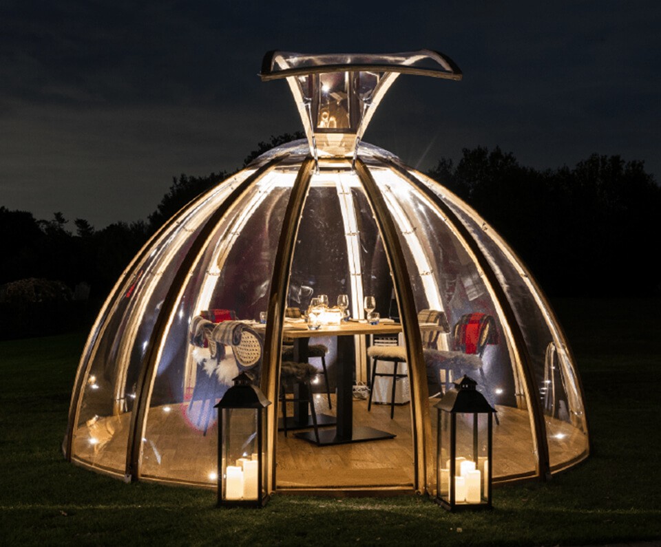 A cozy glass dome tent is dimly lit with fairy lights at night in the gardens of Stirrups Hotel. Inside, a dining table set elegantly for dinner. Two lanterns with candles frame the entrance.